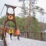 Kaksi retkeilijää kävelee puukantisella sillalla. Maassa on ohut kerros lunta ja taustalla näkyy jäätynyt järvi. Kuva: Jouni Kallio