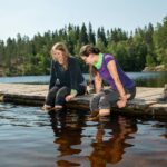 Kaksi retkeilijää istuu laiturilla ja liottaa jalkojaan vedessä. Heidän vieressään laiturilla on vaelluskengät. Kuva: Saara Lavi