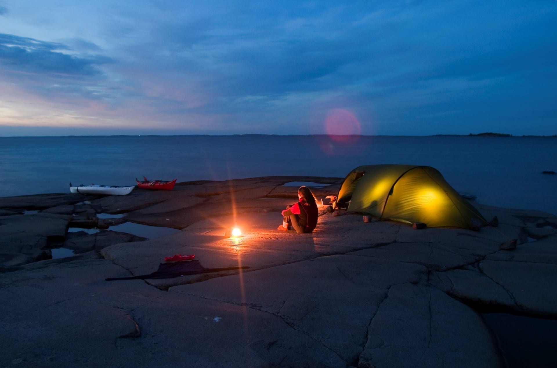 Öinen kuva,  jossa retkeilijä istuu kalliosaarella teltan edessä. Taustalla meri ja rantaan vedettynä kaksi merkikajakkia. Kuva: Markus Sirkka