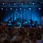 Audience facing an empty stage. On stage there are drums, microphones and an empty bar stool.